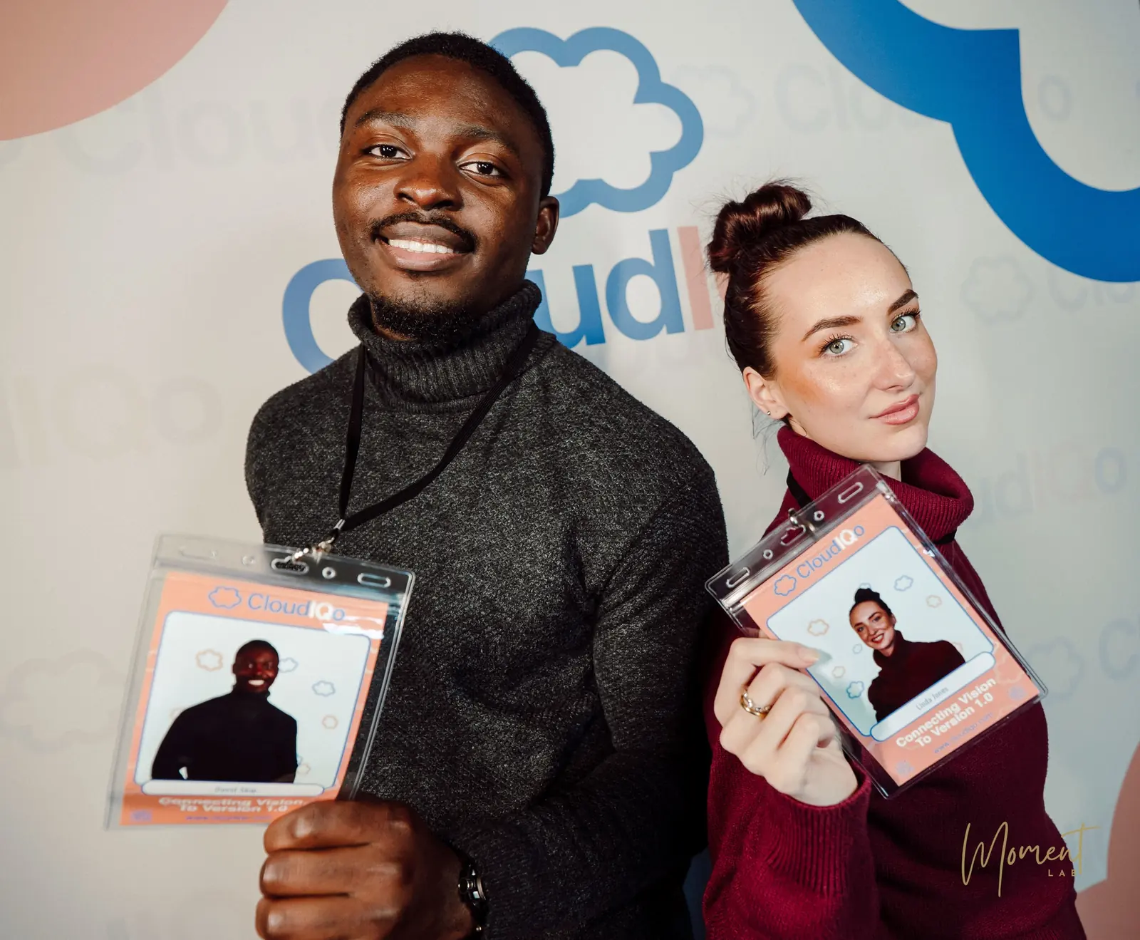 Two attendees holding branded event passes printed with their headshots, in front of the company backdrop