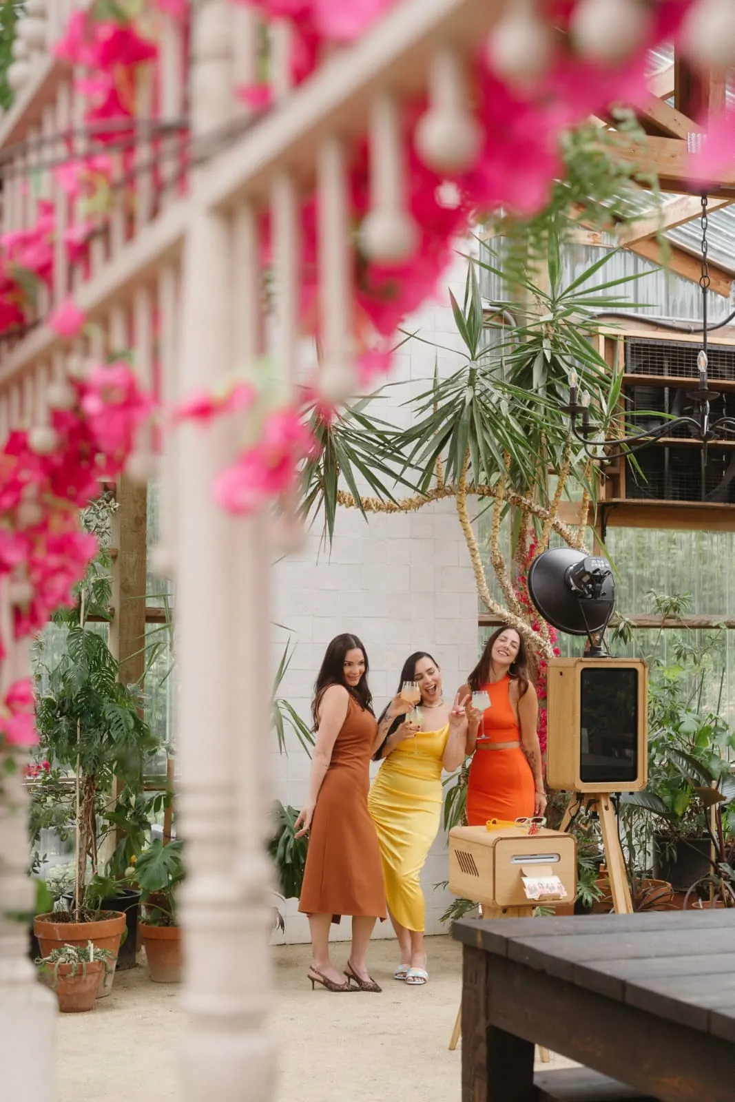 Three guests in bright dresses laughing at the photo booth in a glasshouse venue