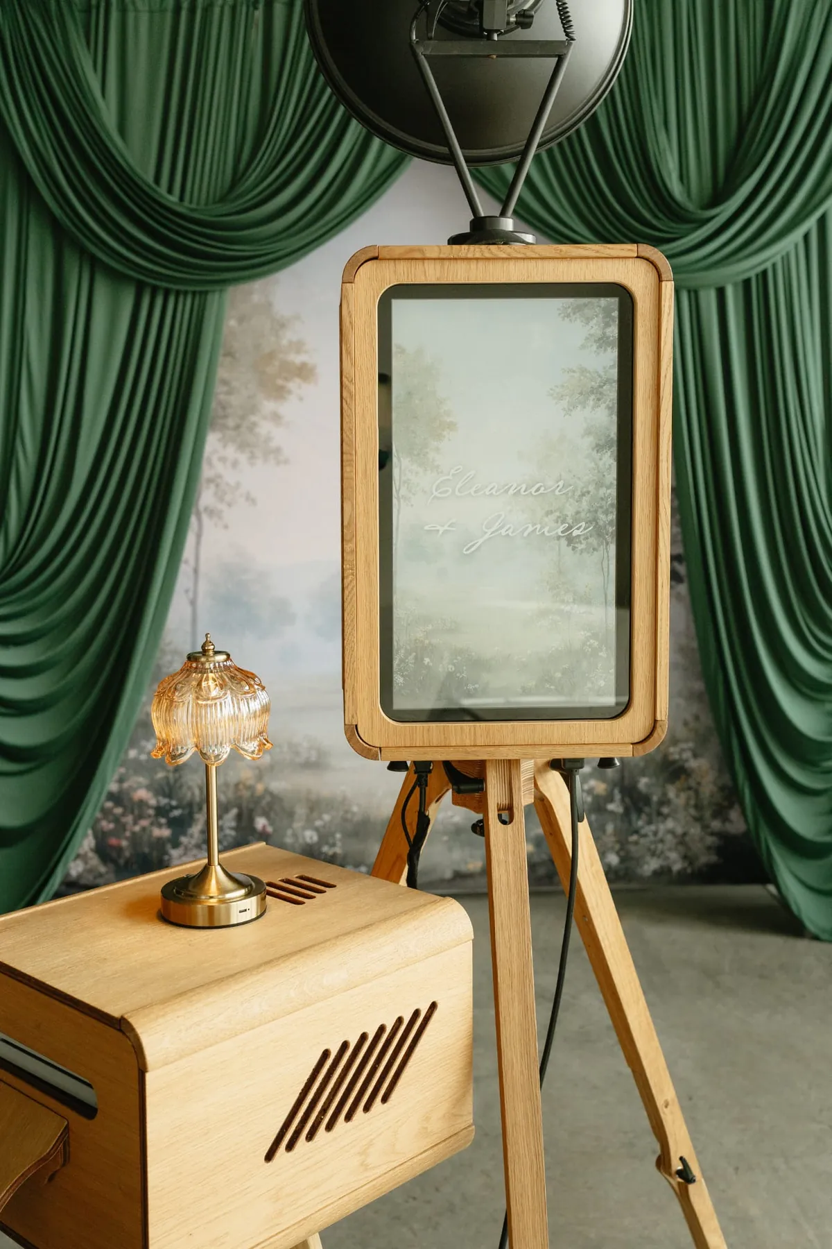 Oak photo booth with a personalised welcome screen, set against green drapes and a painted garden backdrop