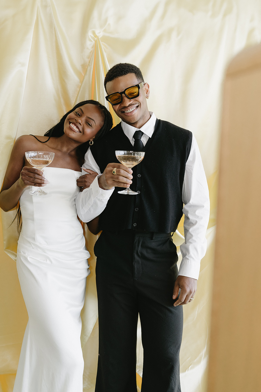 A couple in black-and-white formal attire toasting in front of a champagne satin backdrop