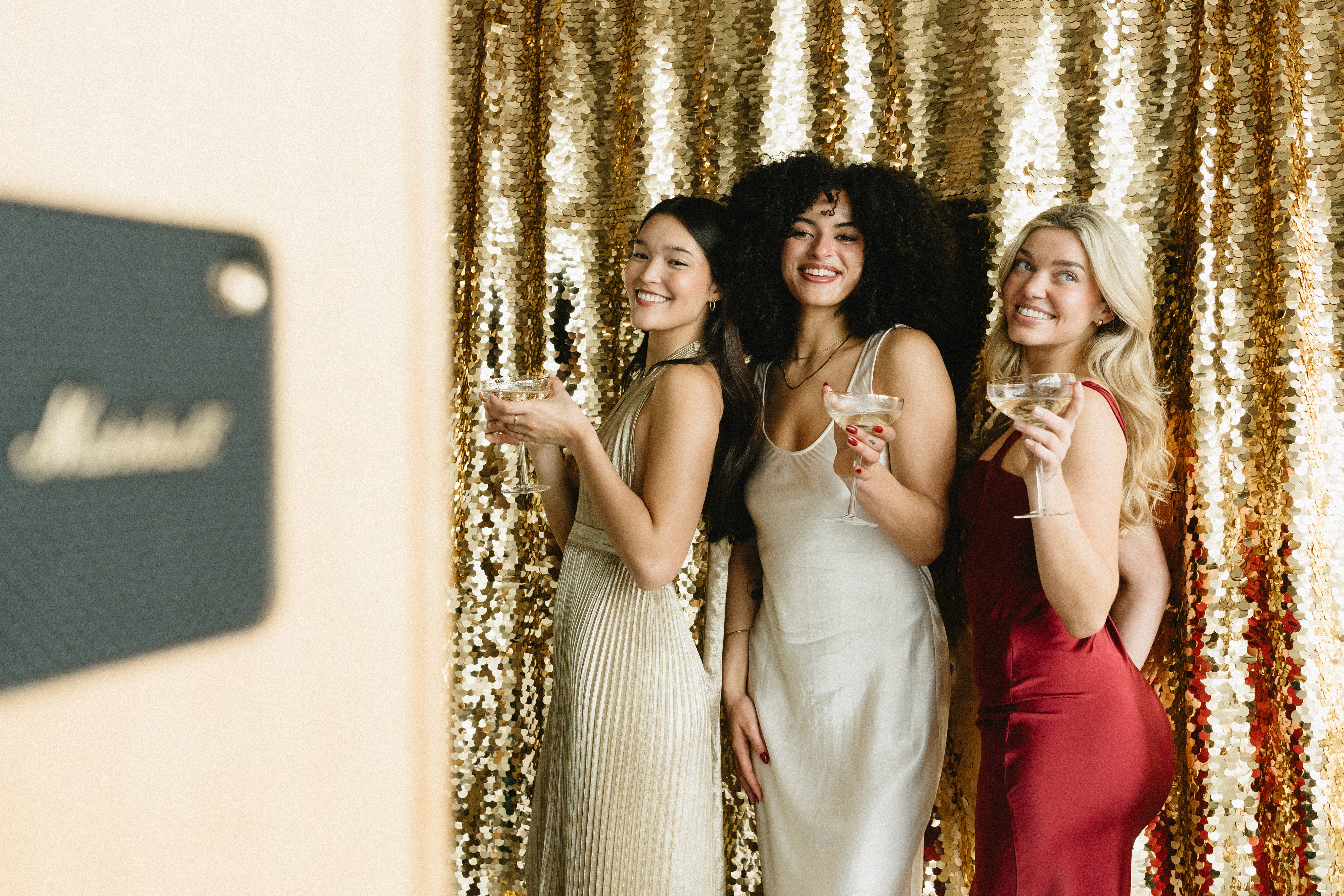 Three guests laughing and posing with champagne coupes in front of a gold sequin photobooth backdrop